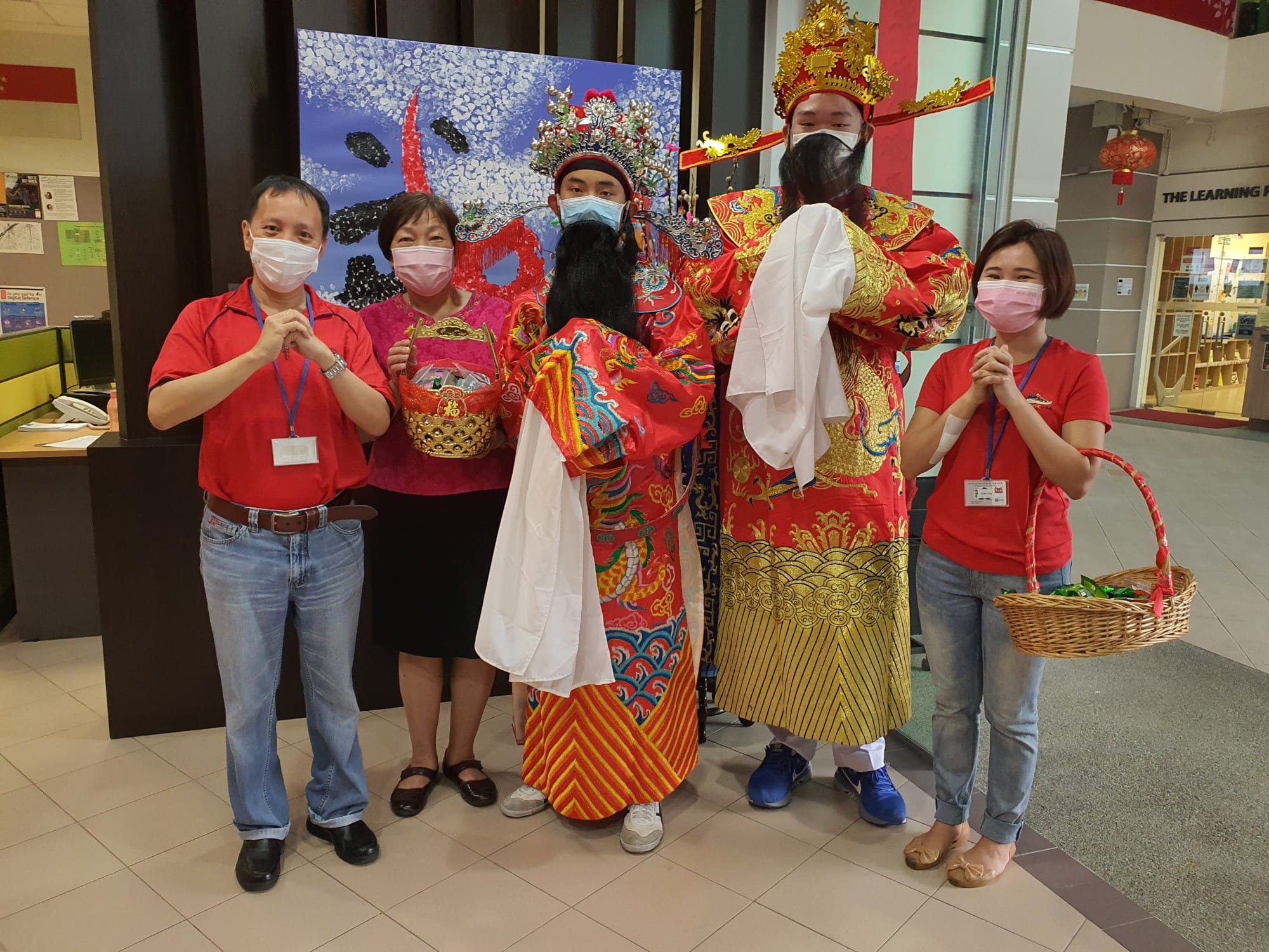 Five people wearing masks pose indoors, two in elaborate traditional Chinese festival costumes and three in red shirts.